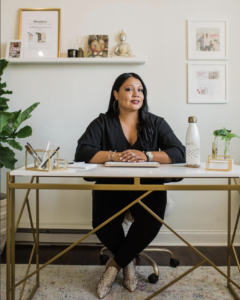 Tina Dixon Spence is a Black woman with long, black hair wearing all black. She is sitting at a tidy, well-decorated desk and looking at the camera.