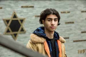 An Iranian teen with ear-length hair parted down the middle is standing in front of a wall with a Star of David on it. He is looking at the camera and is wearing layered hoodies.