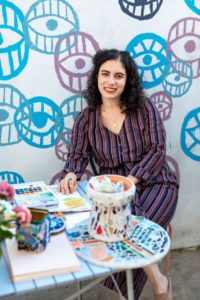 Danielle Abrams is wearing a striped jumpsuit and smiling while sitting at a mosaic table filled with craft supplies. She has dark, curly hair and is wearing red lipstick.