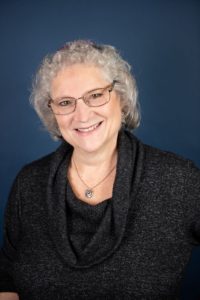 Rabbi Abby Michaleski is a white woman with curly white hair, smiling at the camera. She is wearing glasses, a necklace, and a dark grey sweater.