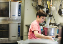 Tova du Plessis is a short, white woman with short hair and bangs wearing a pink t-shirt working in an industrial kitchen, holding a large mixing bowl.