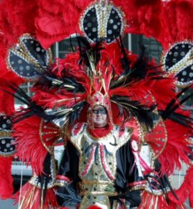 Ed Cox is looking straight at the camera. Black paint is across his eyes, and h is wearing an elaborate costume made of a gold breastplate and black and red feathers that extend from his body.