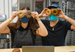 Alexandra (left) is a white woman with long, dark hair wearing an apron next to Jacob Cohen, a white man wearing a hat and black shirt. Both are holding bagels in front of their eyes.