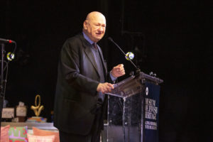 Jack Kliger is a bald, older white man wearing a dark suit. He is standing in front of a microphone and podium on a darkened stage, speaking.