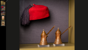 A red fez and two bronze-colored turkish coffee pots are dispalyed in front of a grey-purple background.