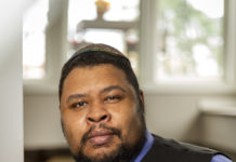 Michael Twitty is a Black man with short, black hair and short beard. He is looking at the camera and wearing a kippah, blue shirt, striped tie and black vest.