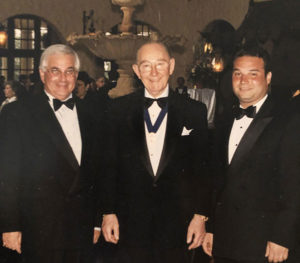Three white men, one middle aged, one older, but younger, stand together in tuxedos smiling for the camera.