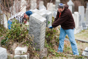 On either side of a large, granite gravestone, two men, both white, trim the hedges between the graves. They are dressed for fall and wearing gardening gloves.