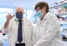 Weissman is a white man wearing a shirt and tie, lab coat and blue mask holding a vial filled with a green liquid. Karikó, a white woman wearing a lab coat with short, brown hair and glasses, also wearing a mask, is looking at the vial.