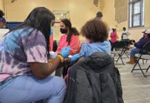A vaccine administrator, a Black woman with long, black hair wearing blue gloves, is giving a shot to a young Black child with curly hair who is pulling up their t-shirt sleeve.