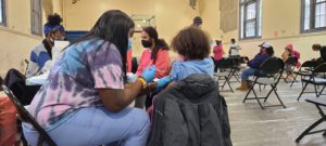 A vaccine administrator, a Black woman with long, black hair wearing blue gloves, is giving a shot to a young Black child with curly hair who is pulling up their t-shirt sleeve.