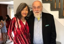 Jacques Lipetz, a bald white man with a white beard wearing a casual suit and using a cane is standing with his arms around Inez Friedman-Lipetz, a woman with long, salt-and-pepper hair wearing a striped pink dress.