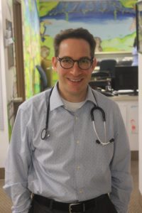 Dr. Craig Barkan is a white man with short, brown hair and thick-rimmed glasses wearing a button-up shirt with a stethoscope around his neck. He has his hands in his pockets and he is smiling at the camera.