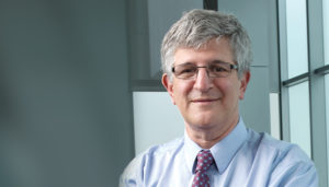 Dr. Paul Offit is a white man with short, grey hair and thin, rectangular glasses. He is wearing a blue button-up shirt and purple patterned tie.