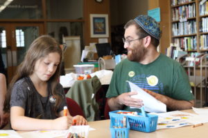 At an arts and crafts table, a white child with long, brown hair is coloring with a market next to an older white person with a thick beard and kippah holding a piece of paper