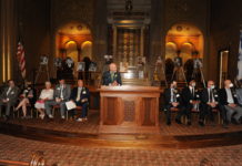 in the Rodeph Shalom sanctuary accented with dark wooden structures, Marc Zumoff, a bald, white man in a suit, is standing at a podium addressing the hall of fame inductees on either side of him.