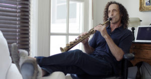 Legs kicked up in front of him, Kenny G, a white man with long, curly, brown hair, is blowing into a soprano saxophone.