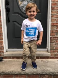 Standing in front of a house's front door, is a small, white child wearing a "Walk Against Hate" white t-shirt, smiling to the camera.