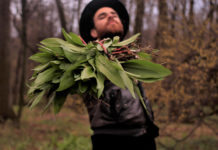 A white man with a brimmed hat and beard has his arm outstretched holding a bouquet of green, leafy ramps.