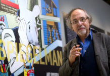 Standing in front of a mural with images from his graphic novel "Maus," Art Spiegelman is holding a pen between his fingers and wearing a blue shirt and cardigan looking at the camera.
