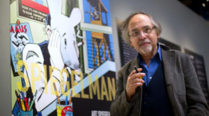Standing in front of a mural with images from his graphic novel "Maus," Art Spiegelman is holding a pen between his fingers and wearing a blue shirt and cardigan looking at the camera.