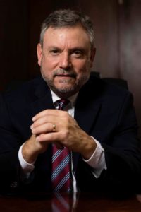 Marc Zucker is a white man with short, grey hair and short beard wearing a suit and striped tie. He is sitting with his hands folded in front of him and looking at the camera.