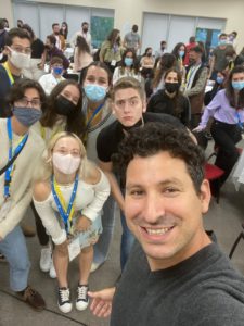 Noam Weissman, a white man with curly, dark hair is smiling and taking a selfie. In the background are dozens of young people, some wearing masks and some making funny faces at the camera.