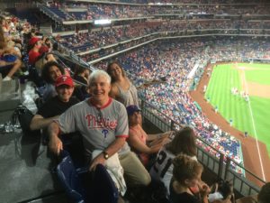 Balcher, a white man with short, white hair is wearing a grey Phillies shirt in the stands about the baseball diamond. To his left are his two sons.