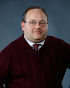 Andre Goretsky is a white man with rectangular glasses and grey hair. He is wearing a dress shirt, tie and sweater and looking at the camera.