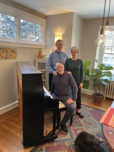 Thom King, a white man in blue sweater, and Elizabeth Shammash, a white woman with white hair tied back, is standing above Zalmen Mlotek, a white man with trimmed, white beard, who is sitting at a piano.