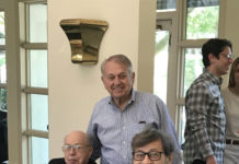 Claude Boni is standing between his sitting brothers at a luncheon table. He is wearing a blue dress shirt and has short, grey hair brushed over to one side. All brothers are smiling.
