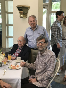 Claude Boni is standing between his sitting brothers at a luncheon table. He is wearing a blue dress shirt and has short, grey hair brushed over to one side. All brothers are smiling.