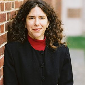 Barbara Mann is a white woman with long, curly hair and dark eyes. She is wearing a dark suit and red turtleneck and standing outside.
