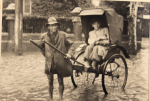In a sepia photo, a young white man is sitting in a rickshaw operated by an older Chinese man wading in water and wearing a wide-brimmed hat.