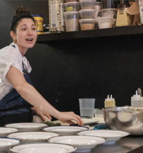 Amanda Shulman, a white woman with dark haired tied back wearing a chef coat and dark apron, is preparing to plate her dish in an array of white bowls laid out in front of her.