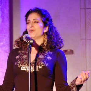Elizabeth Shammash is standing on a stage with purple lighting singing with her arms outstretched in front of a microphone.