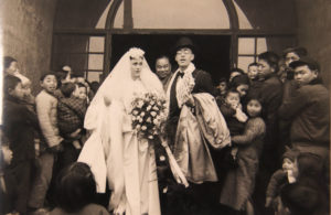 In a sepia photo, Paul Hoffmann in a tuzedo and top hat, is standing beside his bride wearing an ornate white dress and holding a large bouquet in front on a large crowd.