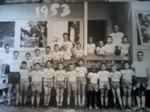 In a black and white photo, young campers in shorts and white t-shirts line up for a picture.