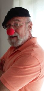 Stuart Goldman is a white man with grey beard. He is wearing an orange shirt, black hat and red clown nose.