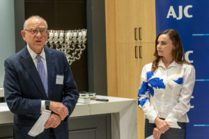 Fred Strober (left) is a white man with thin, white hair and large-rimmed glasses wearing a blue suit and standing next to a white woman in a blue and white dress shirt. They are in front of a menorah.