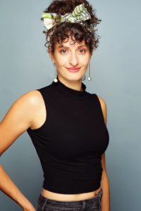 Bex Odorisio is a white woman with curly hair and bangs done up. She is wearing a black sleeveless turtleneck and white dangly earrings.