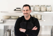 Henry Morgan, standing in a kitchen with his arms crossed, is a white man with short, black hair and mustache wearing all black and smiling.