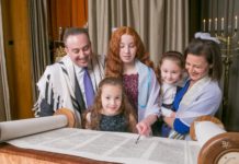 Standing at the bimah, a young white girl with long, red hair reads from the Torah. She is surrounded by her family: a father, mother and two young sisters.