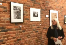 In front of a brick wall lined with framed photographs, Sharon Wohlmuth, a white woman wearing all black with white hair and glasses, stands and looks at the pictures.