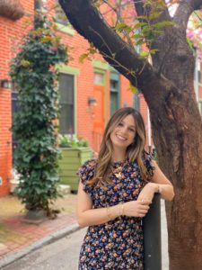 Claudia Kronfeld is a white woman with long brown hair with highlights. She is wearing a floral dress and lots of fold jewelry and is leaning up against a tree.