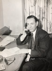 In a black-and-white photo, a white man in a suit sits at a desk with a phone to his ear, looking at the camera.