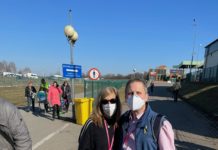 Standing in the middle of the road, Gail Norry, a white woman with blond hair, stands next to Michael Balaban, a white man with short, grey hair. Both are wearing N95 masks.