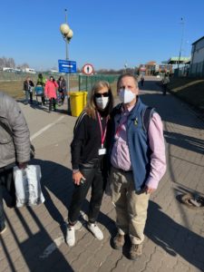 Standing in the middle of the road, Gail Norry, a white woman with blond hair, stands next to Michael Balaban, a white man with short, grey hair. Both are wearing N95 masks.