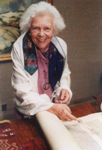 Dressed in a tallit and with a yad on an open Torah, Judith Kaplan Eisenstein, a white woman with curly, white hair smiles at the camera.