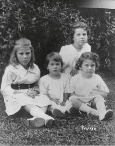 In a black-and-white photo, four young white girls in white dresses sit outside on the grass.
